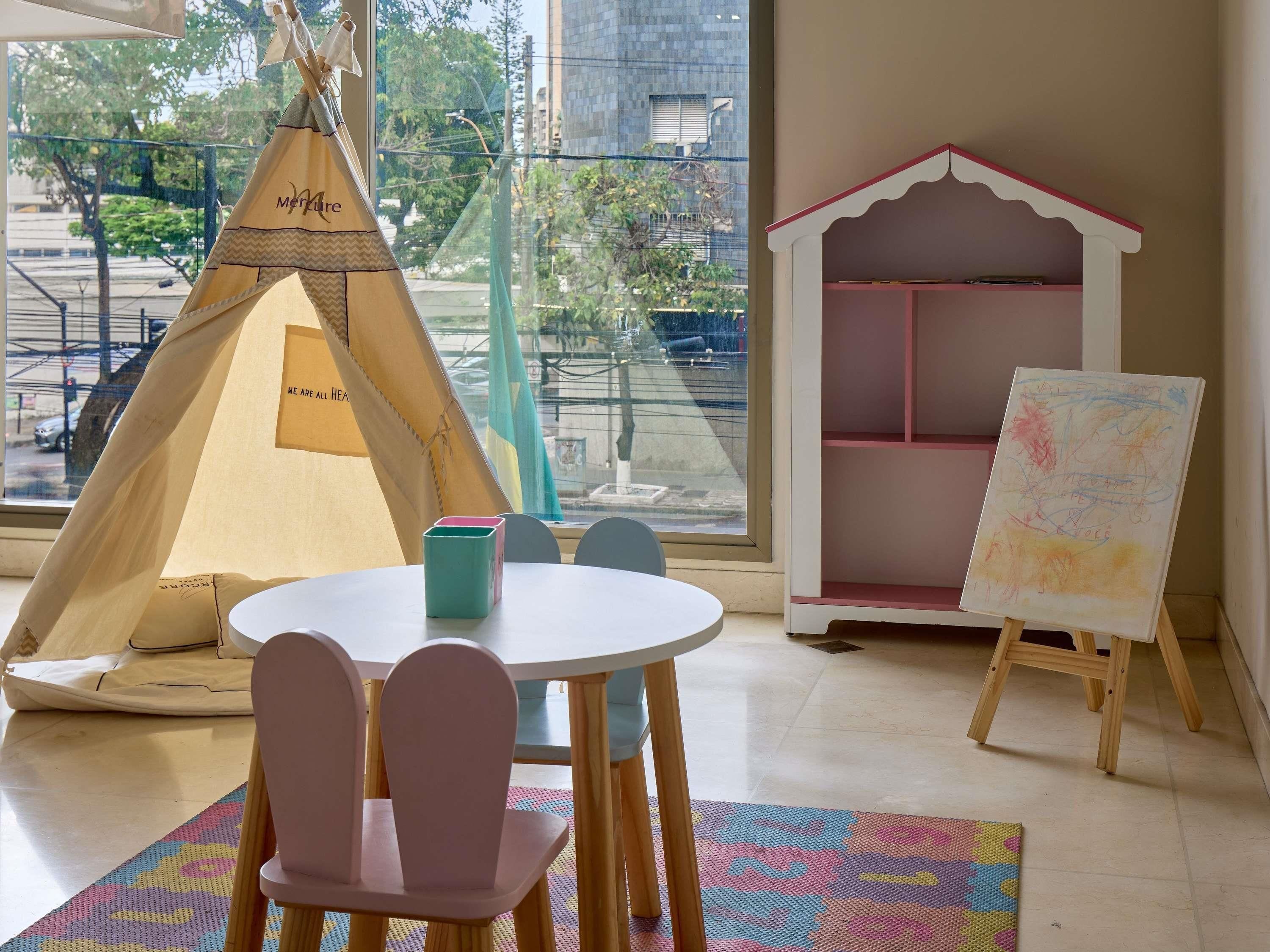 Una habitación con una mesa blanca, una silla rosa, un tapete colorido, una tienda de campaña y una casita de juegos con un cuadro.