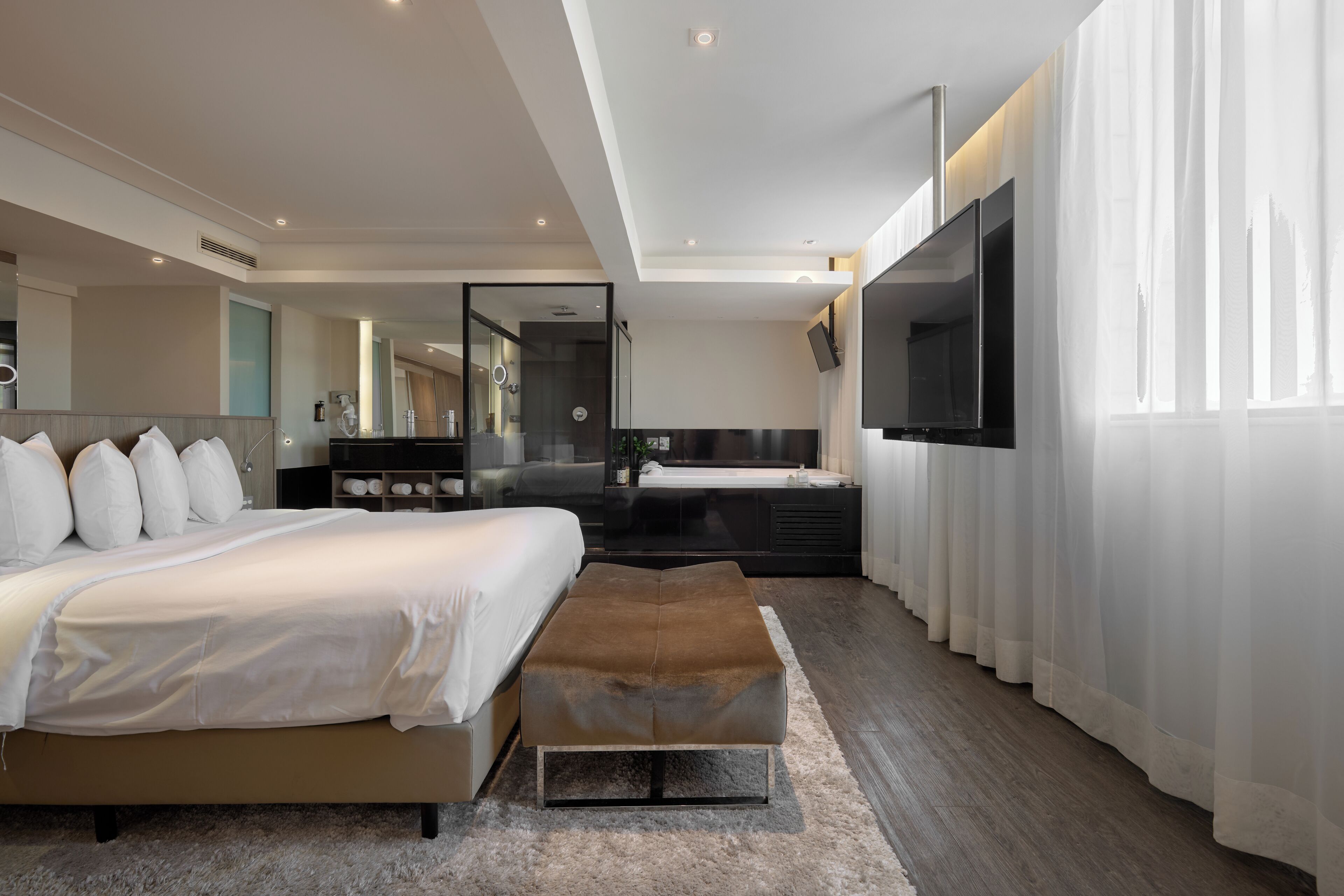 A modern bedroom with a large bed, a TV, a bathroom, and a view of the city.