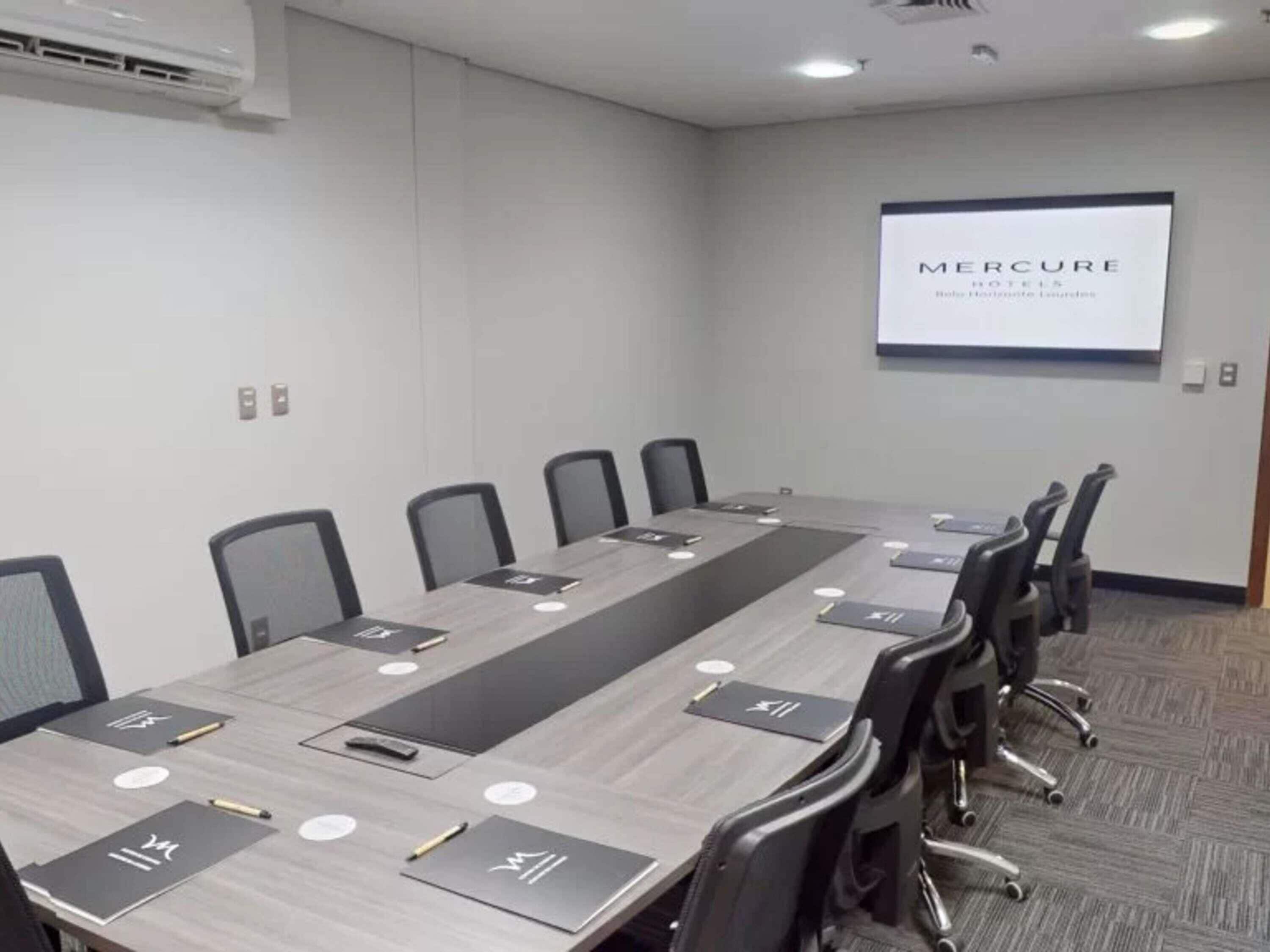 Un salón de conferencias con una larga mesa, sillas, tarjetas de nombre y una pantalla de proyector mostrando 'Mercure Hotels'.