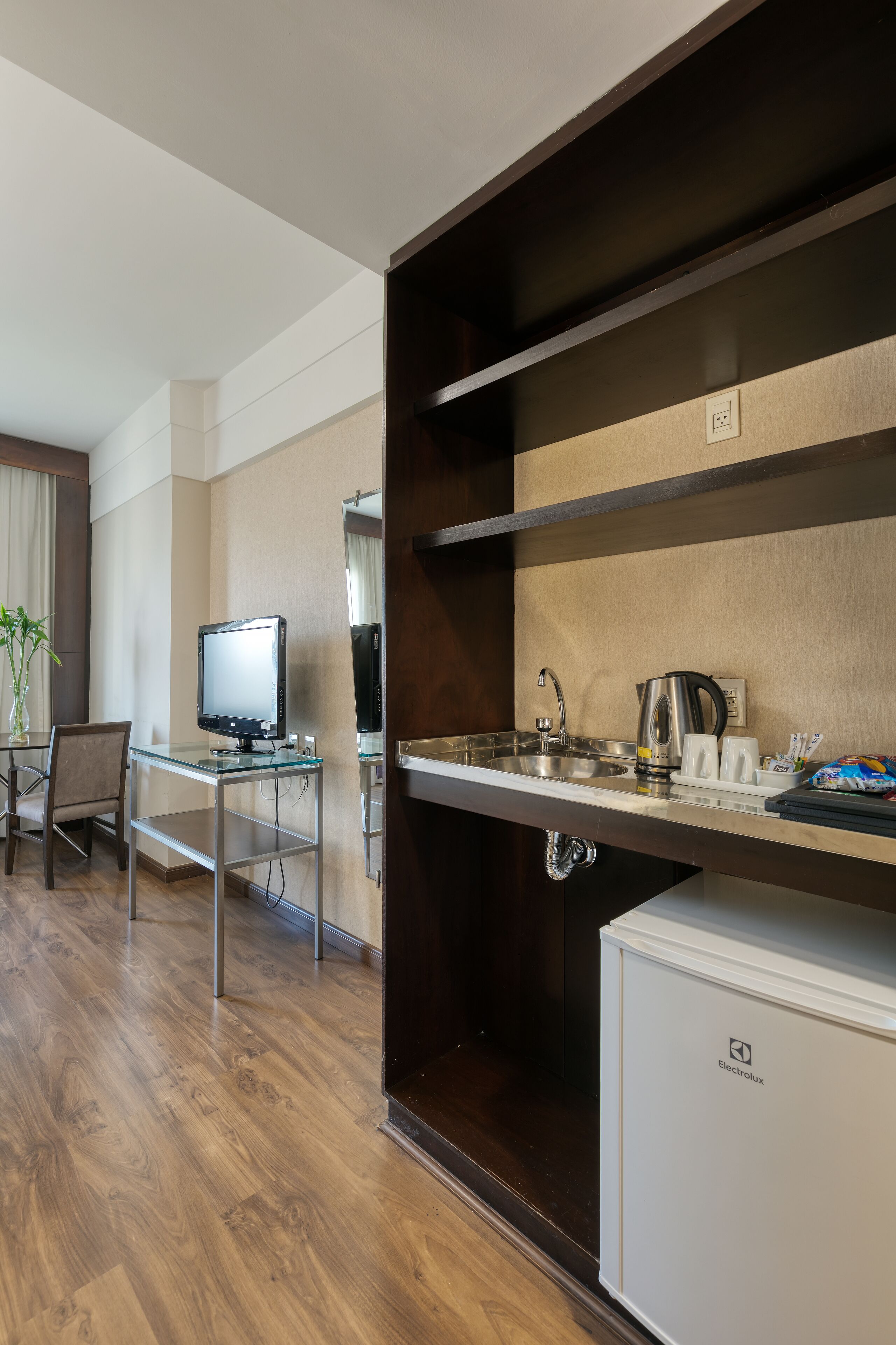 Une chambre d’hôtel avec un plancher en bois, un bureau en verre avec un ordinateur, un petit réfrigérateur et un coin cuisine équipé d’une bouilloire et d’une machine à café.