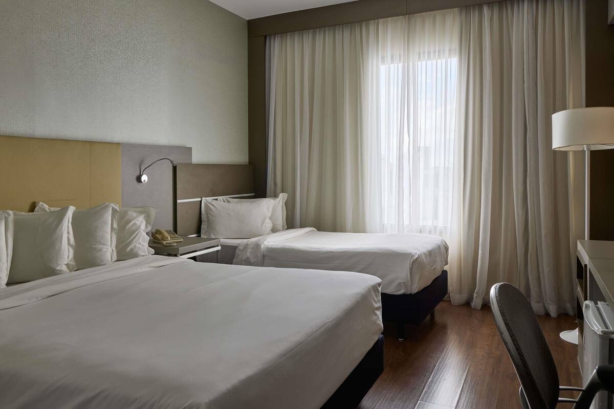 A hotel room with two beds, a desk, a chair, a lamp, and a large window with curtains.