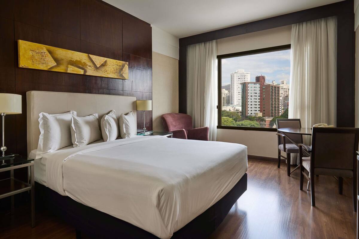 A hotel room with a large bed, a desk, a chair, a lamp, and a view of the cityscape.