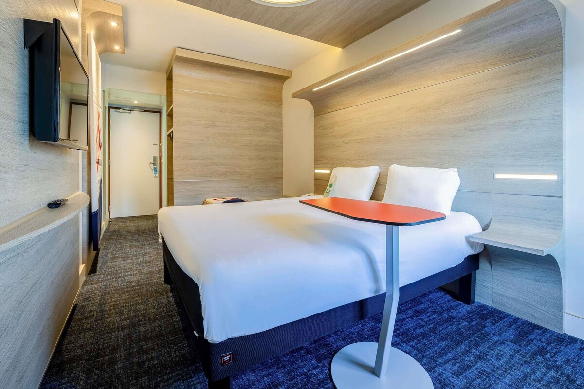 A modern hotel room with a bed, a TV mounted on the wall, and a carpeted floor.