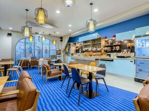 Un restaurant amb un terra de catifes blaves a ratlles, taules i cadires de fusta, llums penjants i una zona de cuina amb prestatges que mostren aliments.