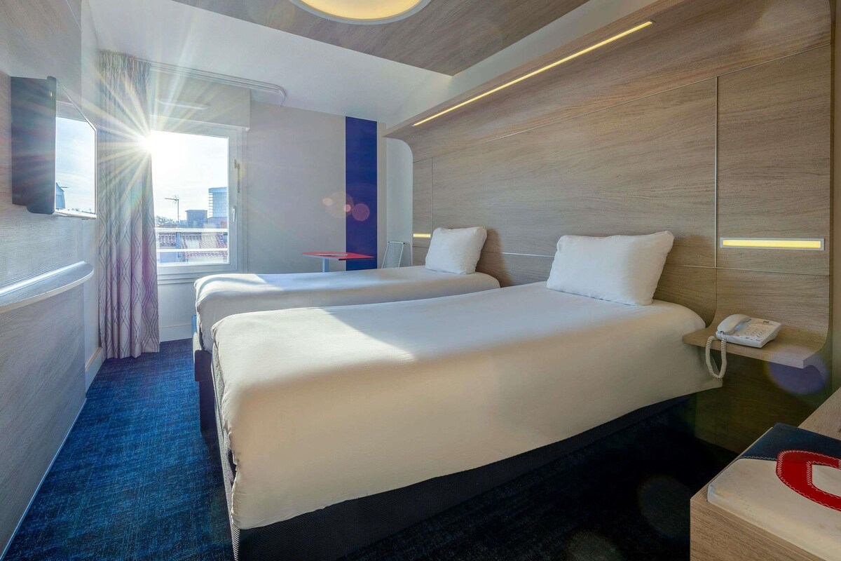 A hotel room with two beds, a desk with a phone, and a view of the city.