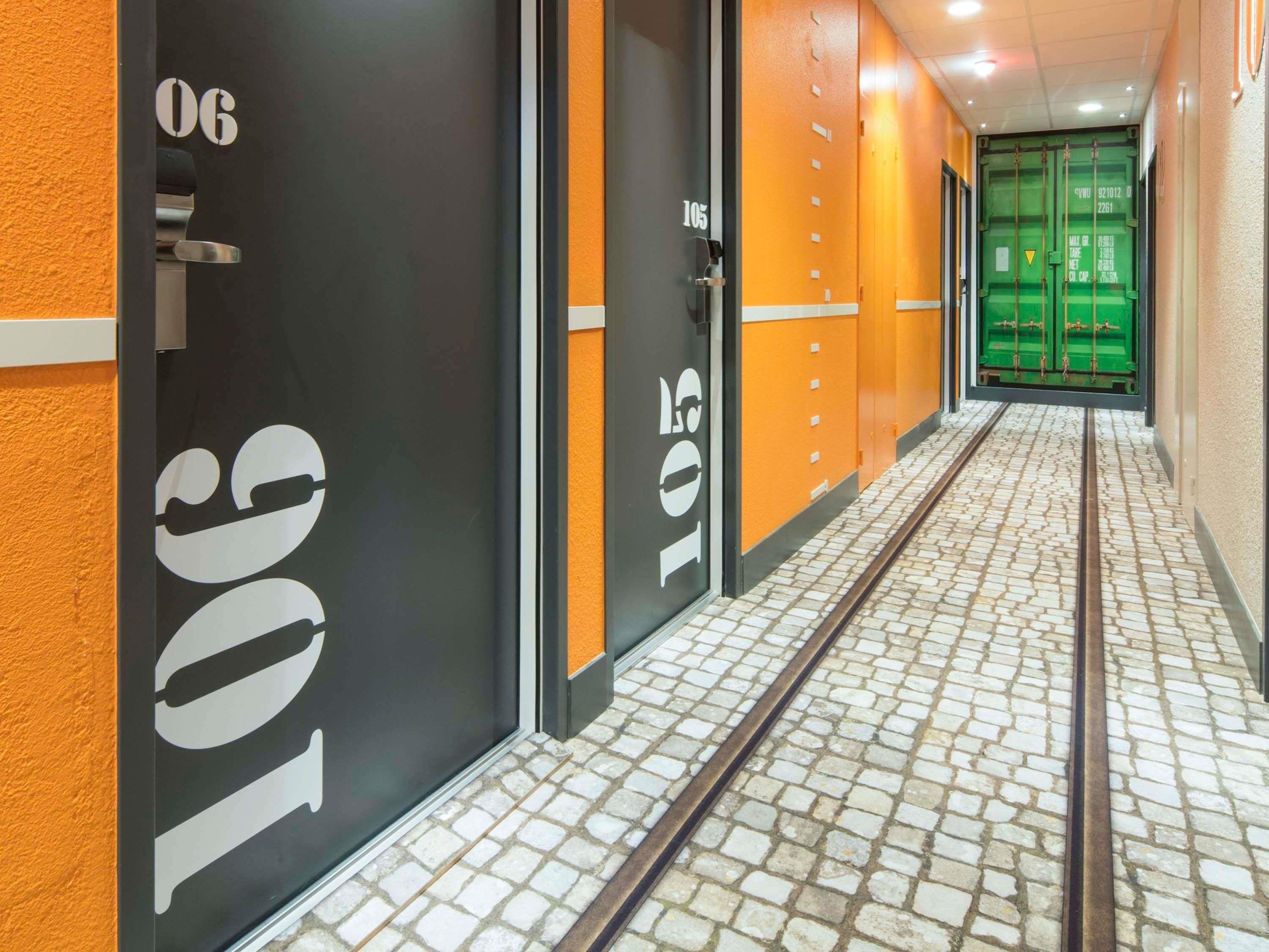 A corridor with numbered doors, a tiled floor, and green shipping containers in the background.