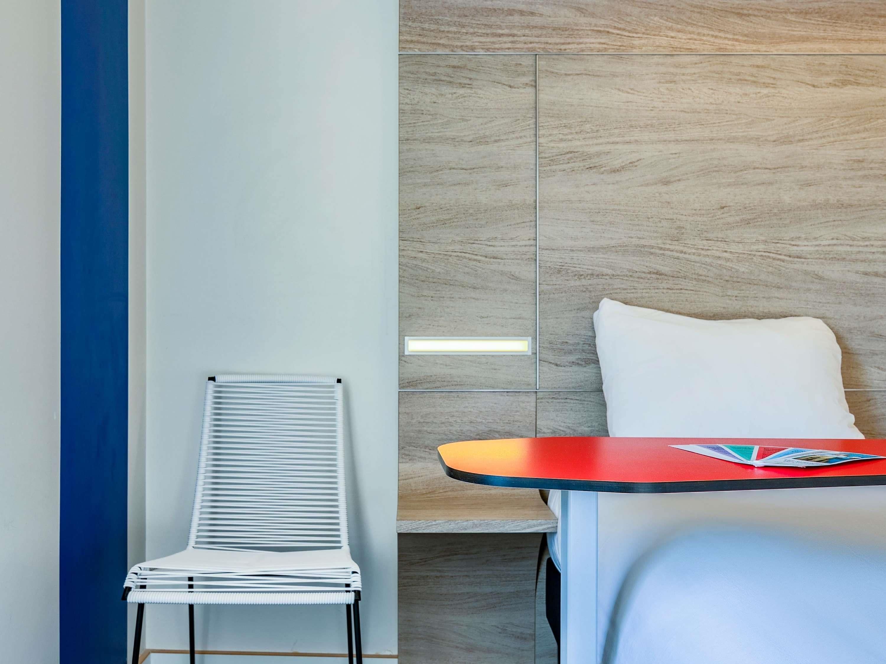 Una habitación de hotel moderna con una silla blanca, una mesita con una revista y una pared de acento roja.