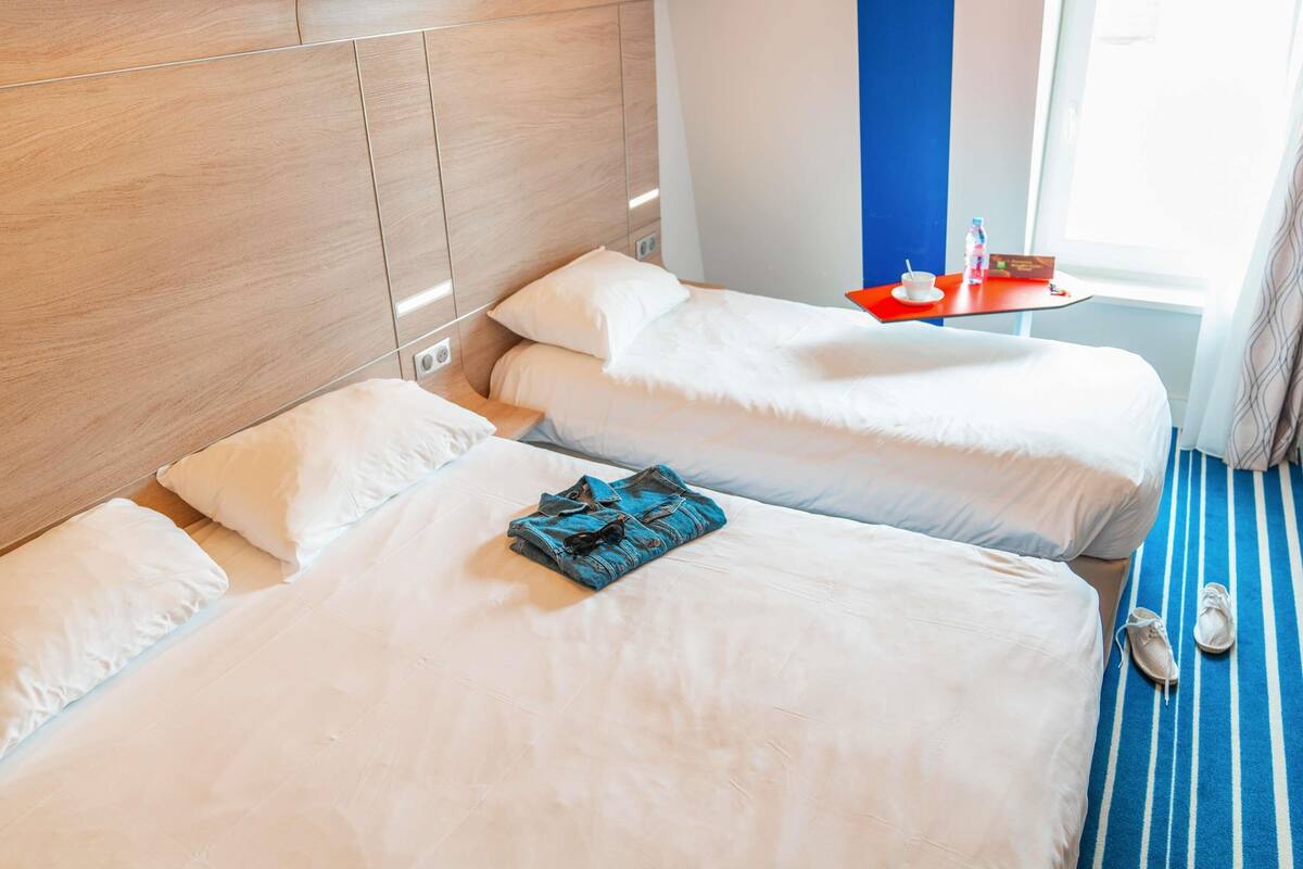 A hotel room with two beds, a wooden headboard, a red table with a cup and saucer, and a striped carpet.
