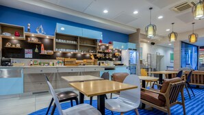 A modern cafe with wooden tables and chairs, a counter with various items, and a blue patterned floor. - ibis Styles La Rochelle Centre Vieux Port (La Rochelle)