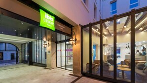 Exterior de um hotel ibis Styles com entrada de vidro, uma placa de identificação e uma área de estar visível pelo vidro.
