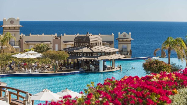 Zona de piscina en un resort con vistas al mar, un pabellón y mobiliario exterior.