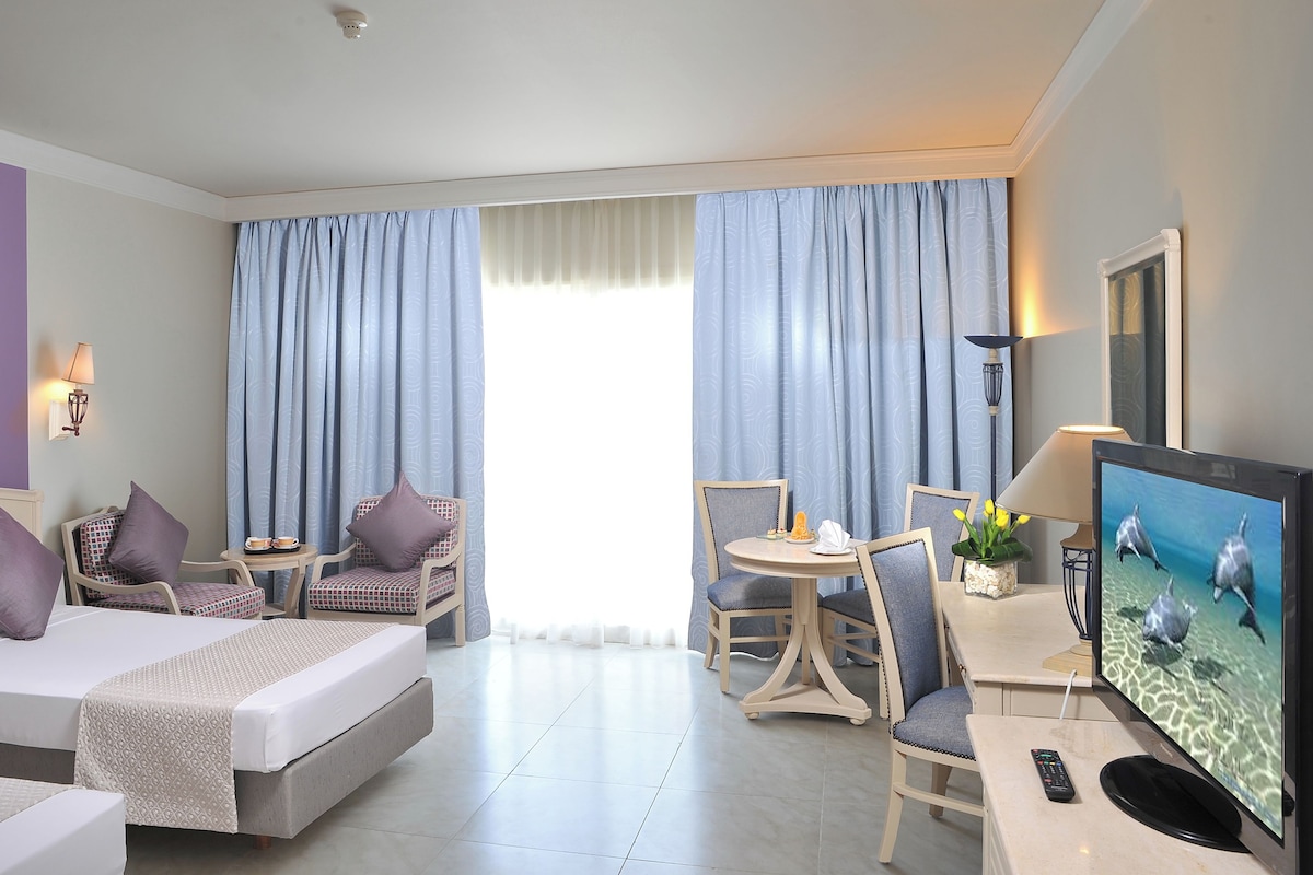 Una habitación de hotel con una cama, dos sillas, una mesita, un televisor y una lámpara.