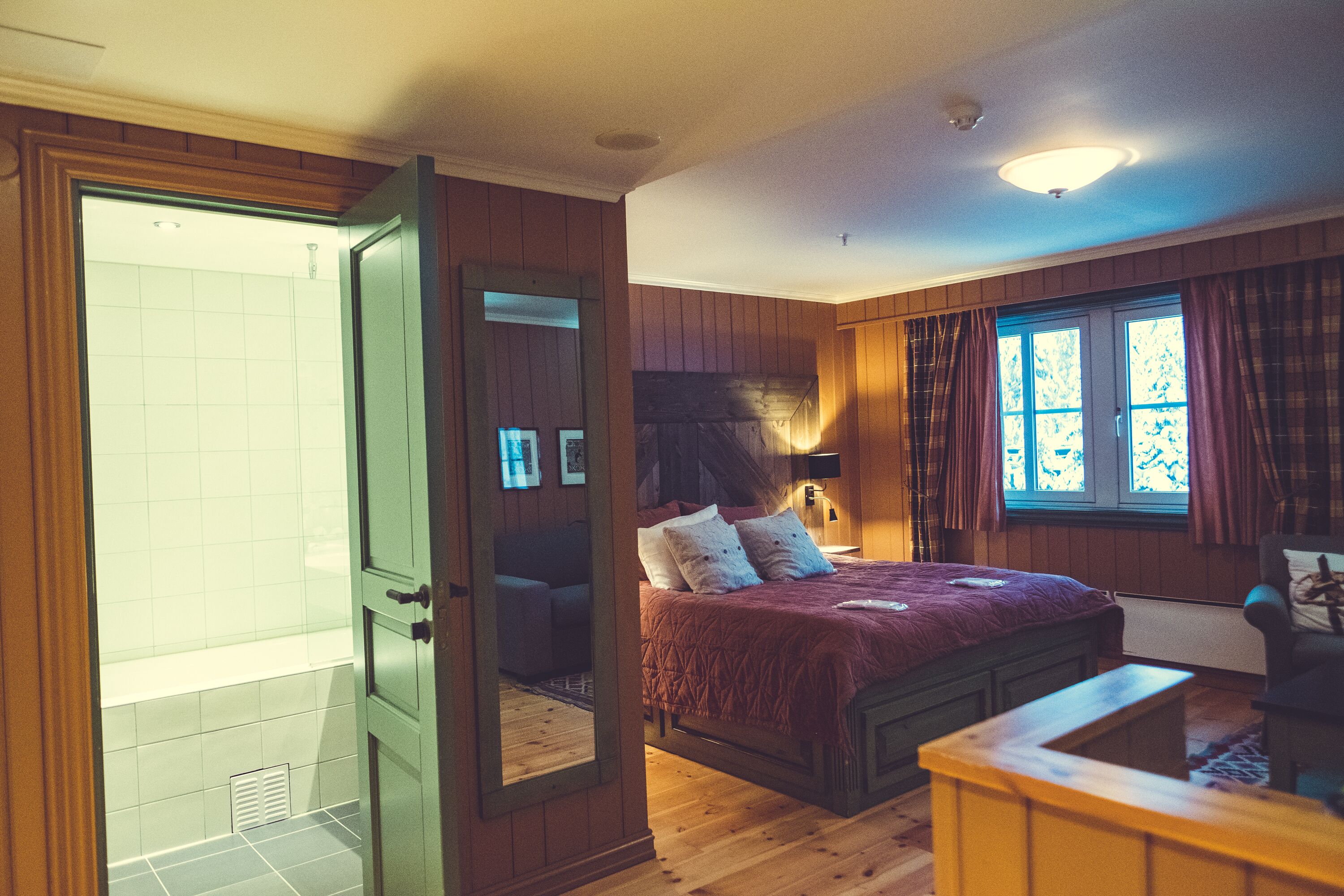A bedroom with a wooden bed, a nightstand, a chair, a window with curtains, and a door leading to a bathroom.