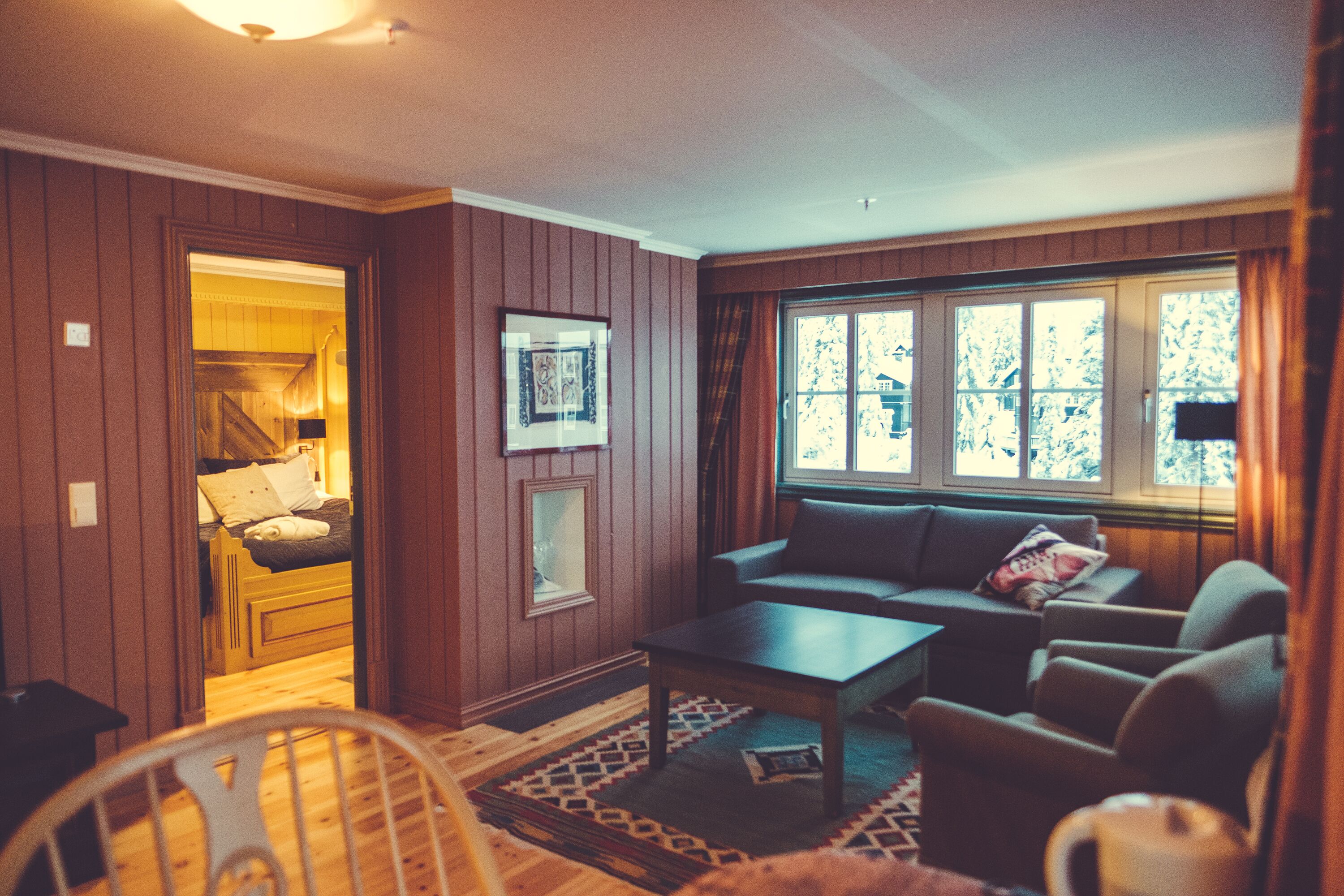 Una sala de estar acogedora con un sofá, una mesa de centro y una silla. Se ve una ventana con cortinas, una puerta que conduce a otra habitación y un cuadro en la pared.