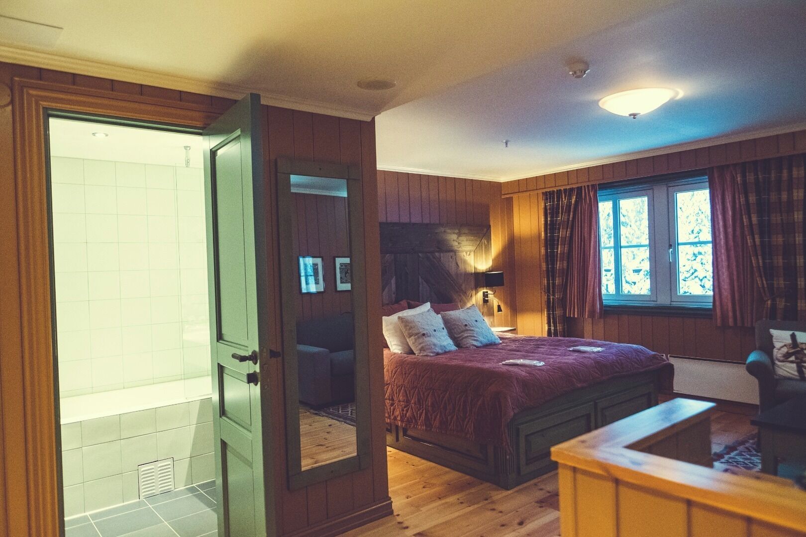 A bedroom with a bed, a wooden headboard, a nightstand, a chair, a window with curtains, and a door leading to a bathroom.