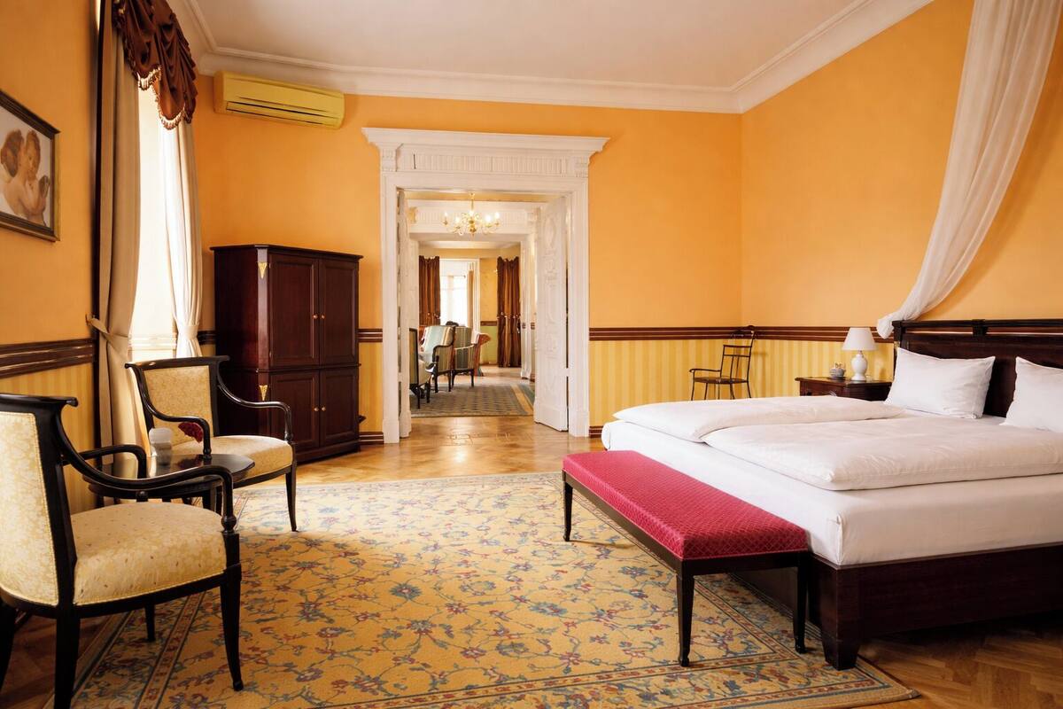 Habitación de hotel con una cama grande, un armario de madera, una zona de comedor con sillas y un pasillo que conduce a otra habitación.