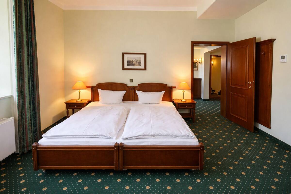 Habitación de hotel con una cama grande, dos mesitas de noche con lámparas, un cuadro enmarcado en la pared y una puerta que da a otra habitación.