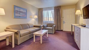 A hotel room with a sofa, two armchairs, a small table, a desk with a lamp, a TV, and a door. - Three Rivers Inn (Biggs Junction)