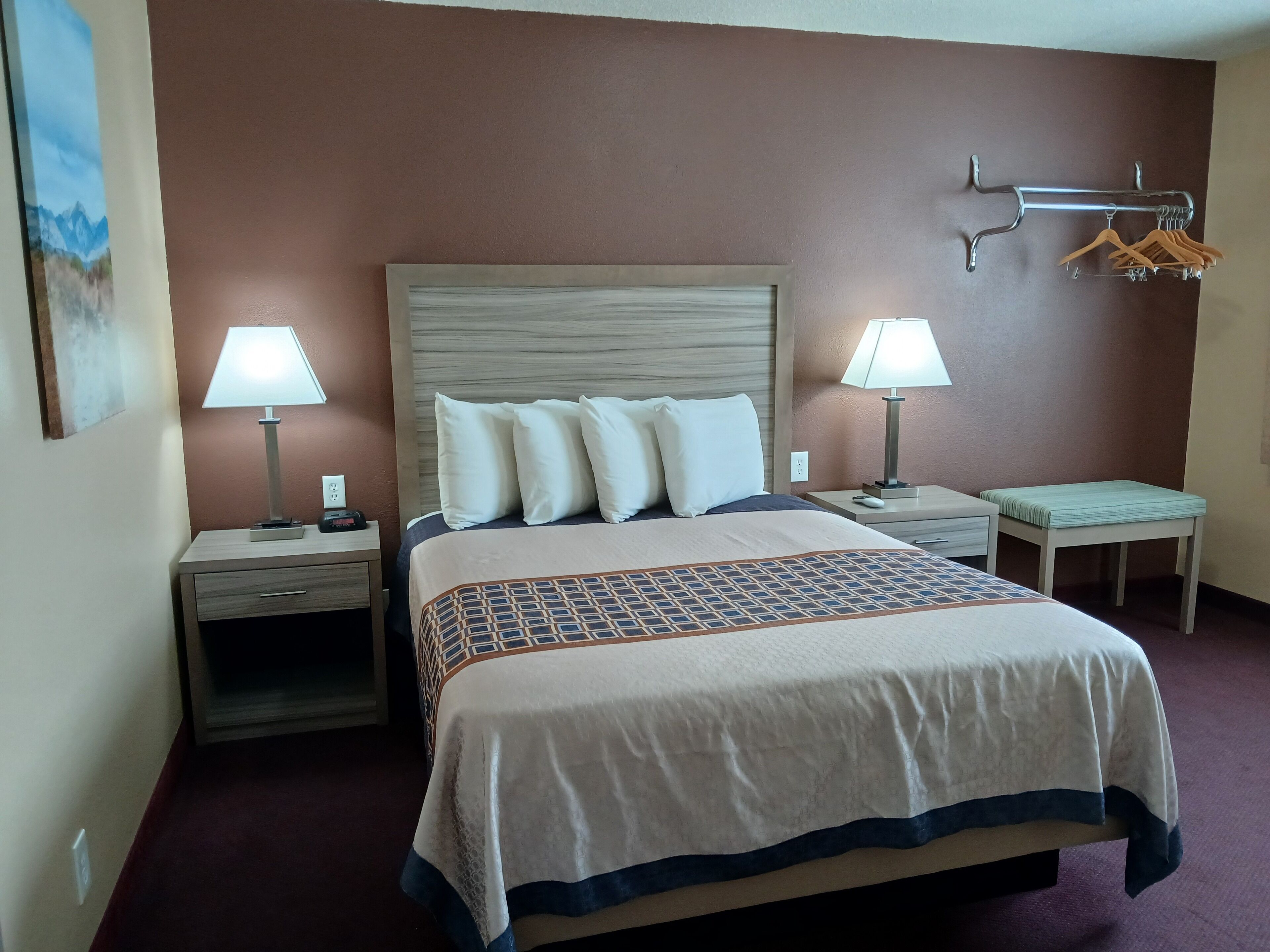 A hotel room with a large bed, two bedside tables with lamps, a wall-mounted coat rack, and a small table with a cushion.