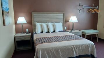 A hotel room with a large bed, two bedside tables with lamps, a wall-mounted coat rack, and a small table with a cushion.