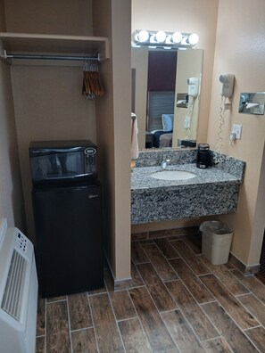 A compact bathroom with a black refrigerator, a sink with a granite countertop, a mirror with lights, and a hairdryer on the wall.