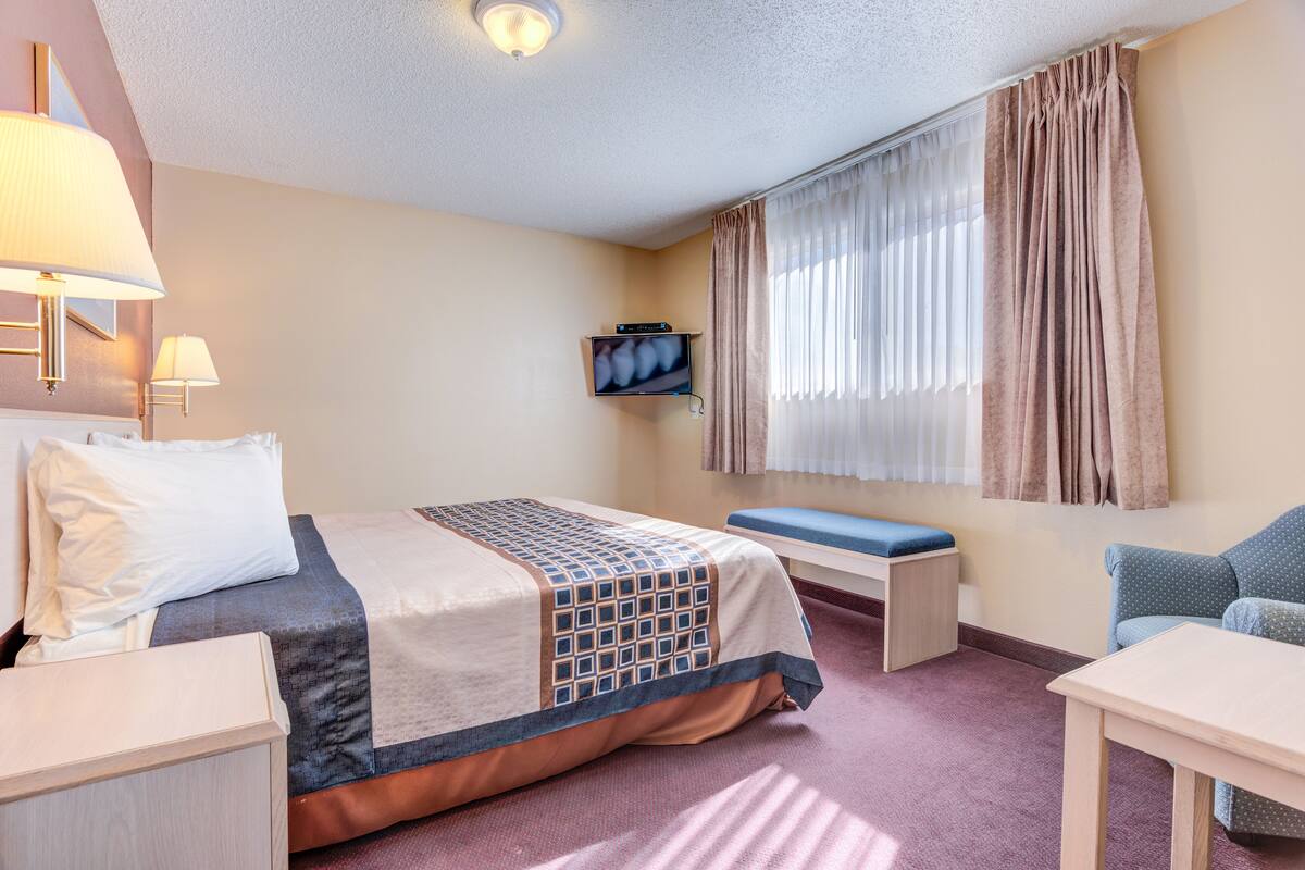 A hotel room with a bed, bedside table, lamp, TV, window with curtains, and a small table.