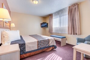 A hotel room with a bed, bedside table, lamp, TV, window with curtains, and a small table.