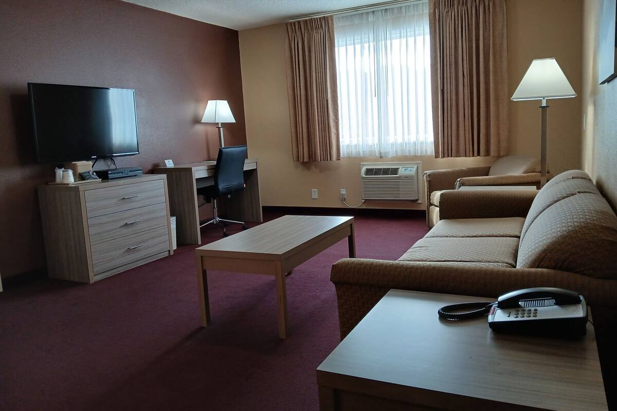 A hotel room with a flat-screen TV, a desk with a chair, a sofa, a coffee table, a telephone, and an air conditioning unit.