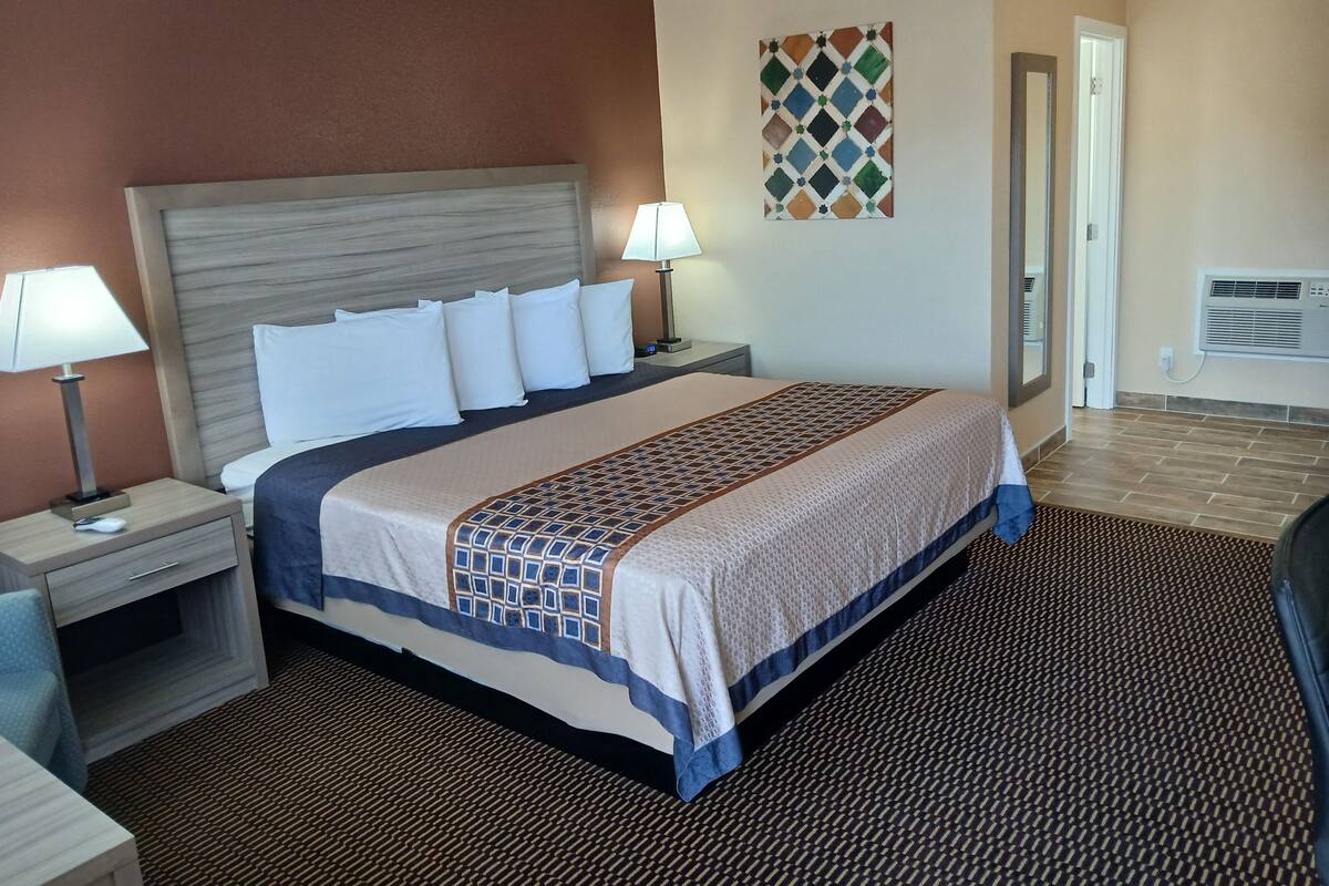 A hotel room with a bed, two bedside tables, a lamp, a wall decoration, and an air conditioning unit.