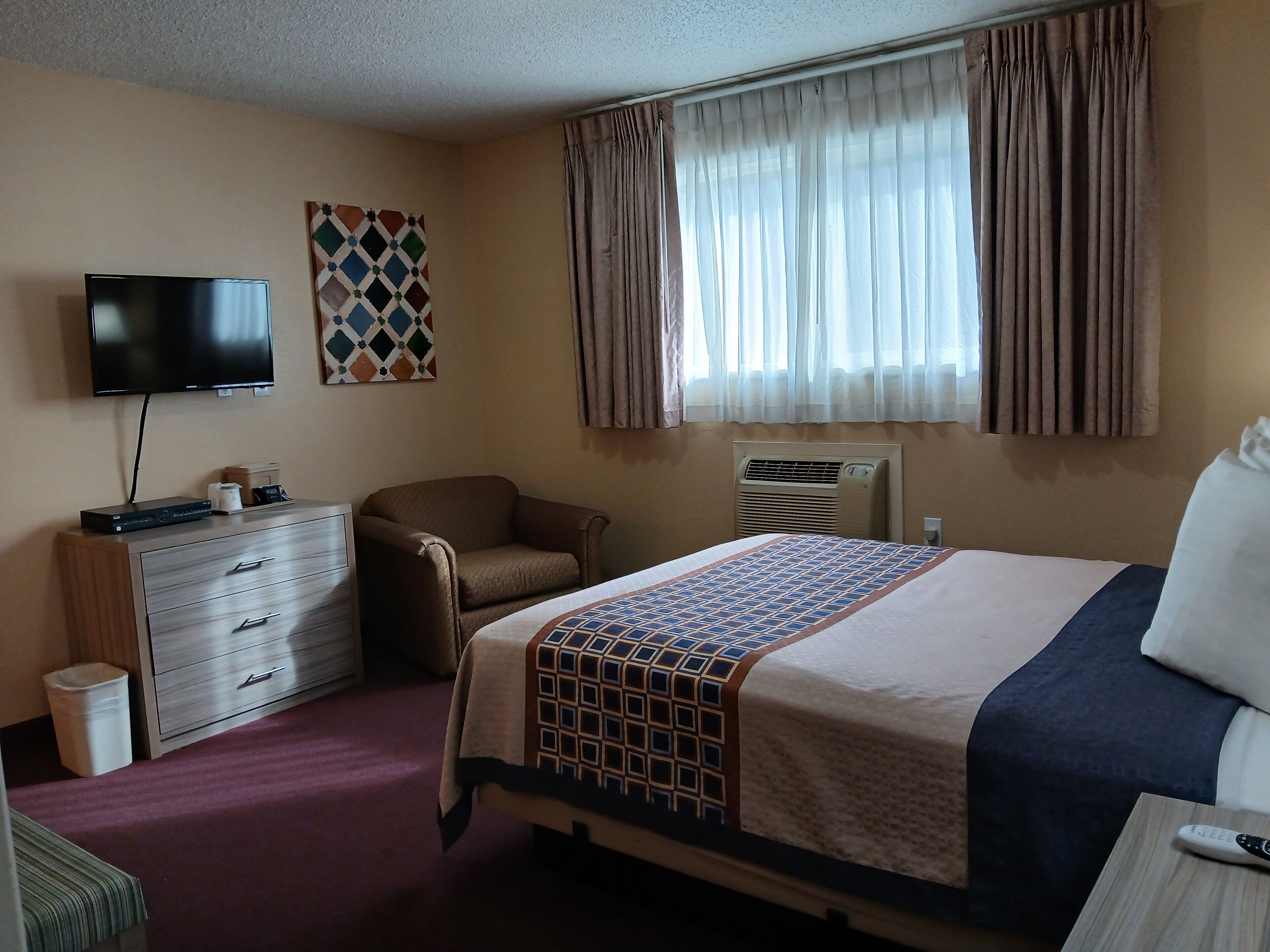 A hotel room with a bed, a TV, a desk, a chair, a dresser, and a window with curtains.