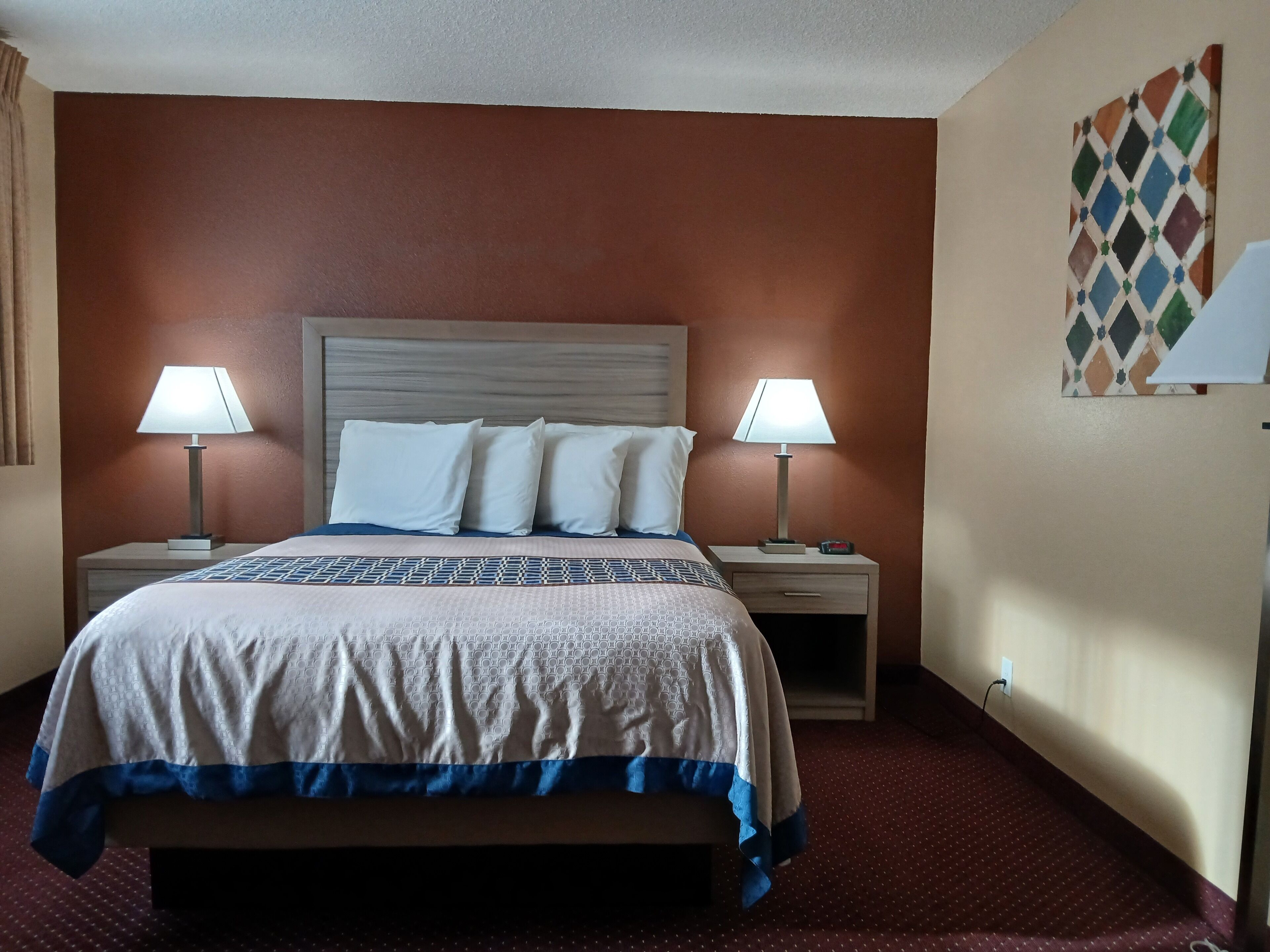 A hotel room with a large bed, two bedside tables with lamps, a patterned bedspread, and a wall with a geometric artwork.
