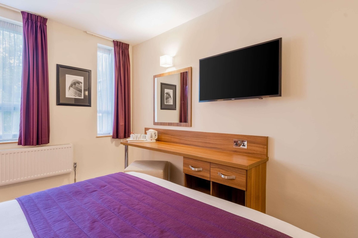 A hotel room with a bed, a wooden desk, a television, and two framed pictures on the wall.