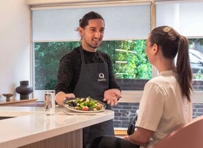 A person in a black apron with "Quality" logo serving a salad to a seated individual in a restaurant with a brick wall and greenery visible through the window. - Quality Hotel Hampstead (London)