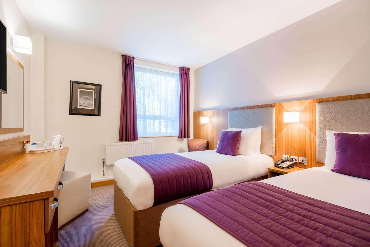 Quarto de hotel com duas camas, uma mesa, uma cadeira, janela com cortinas, um quadro emoldurado e uma mesinha de cabeceira com telefone.