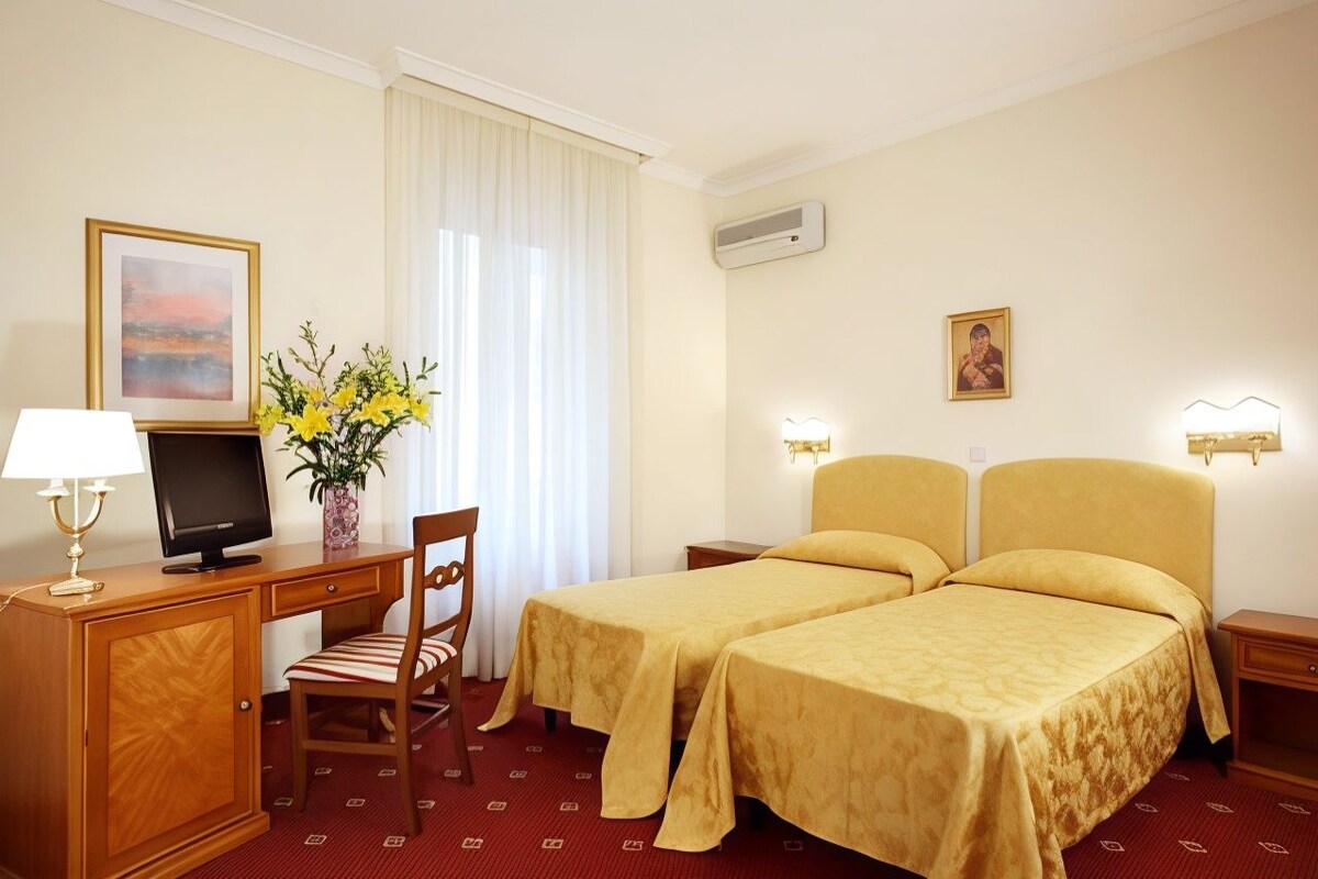 A hotel room with a desk, two beds, a chair, a TV, a vase with flowers, and wall-mounted lights.