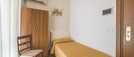 A small, neatly arranged bedroom with a single bed, a wooden wardrobe, a chair, and a wall-mounted air conditioner.