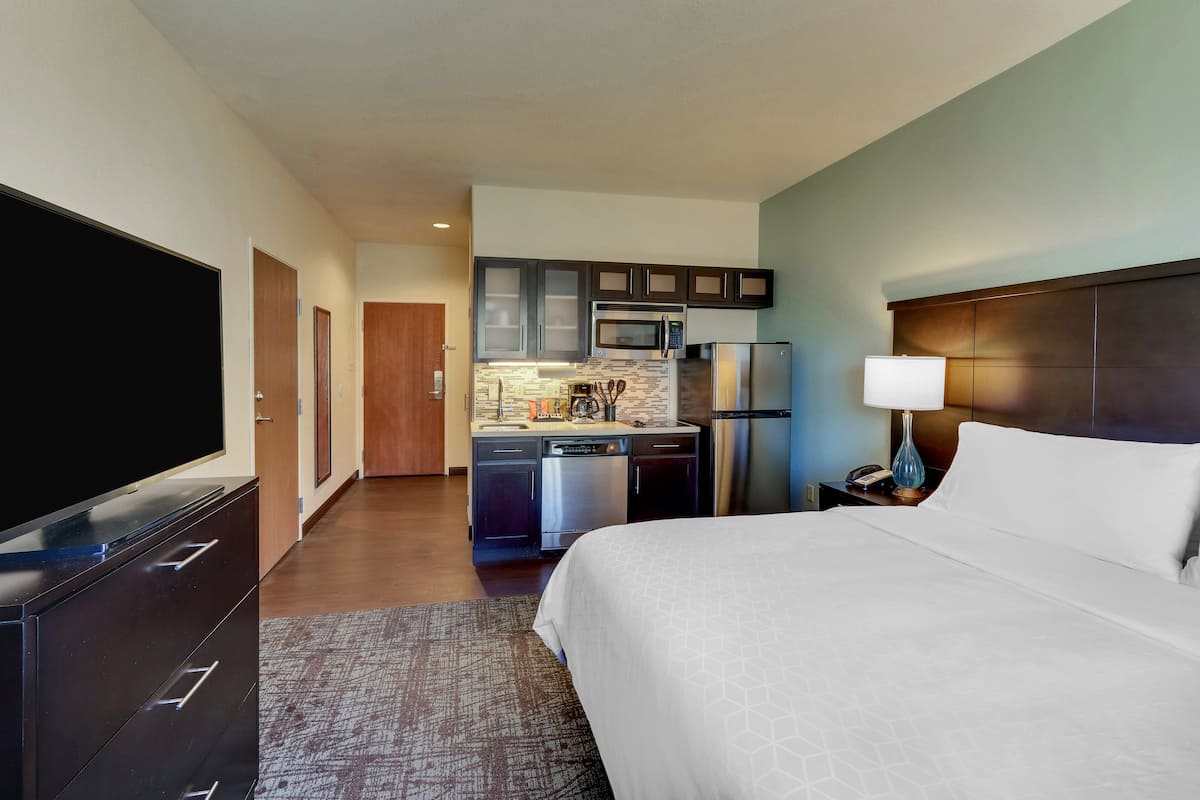A hotel room with a large bed, a TV, a kitchenette with a microwave, refrigerator, and cabinets, and a nightstand with a lamp.