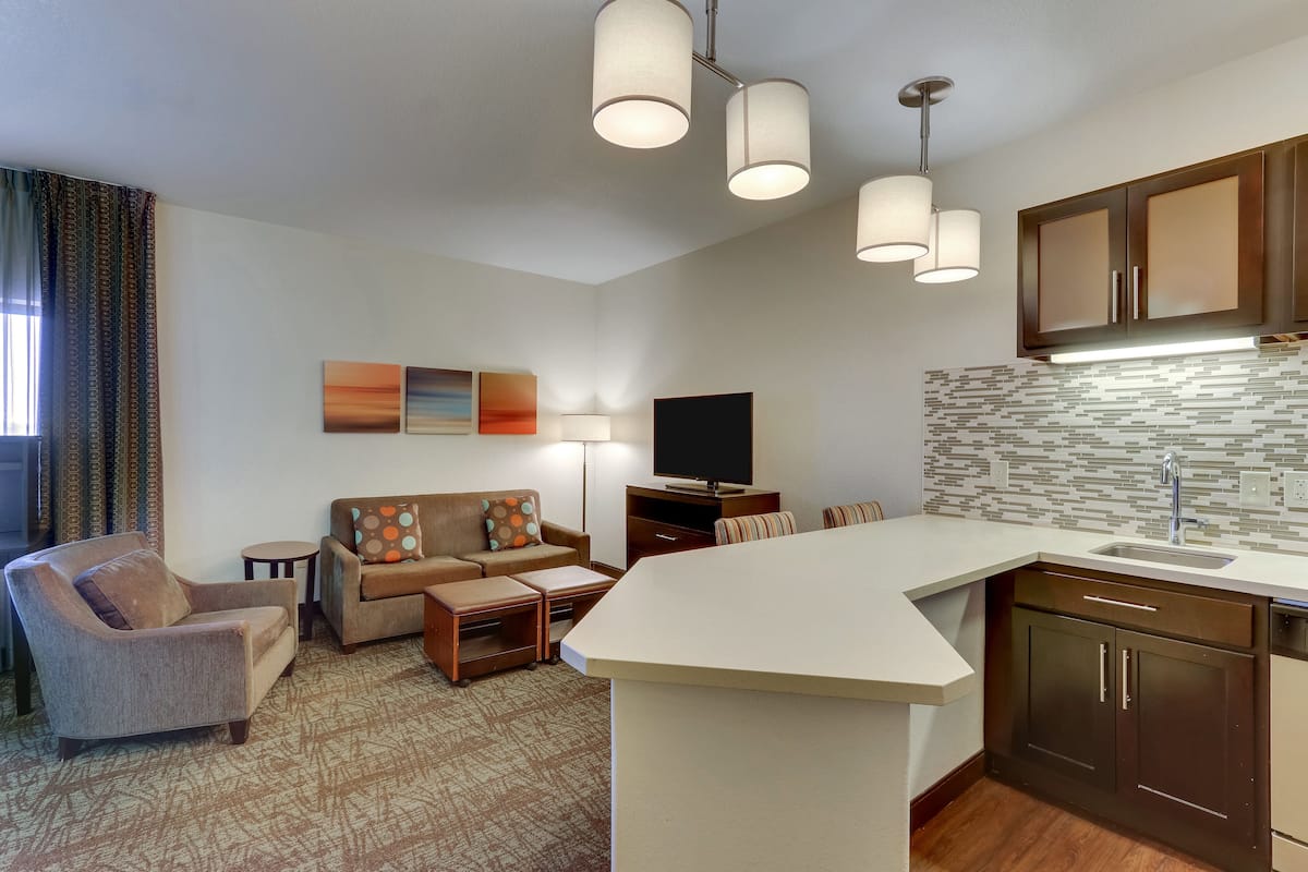 A modern living room with a sofa, armchair, and a flat-screen TV. There is a kitchen area with a sink and cabinets.