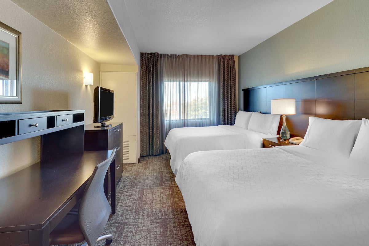A hotel room with two beds, a desk with a TV, a lamp, and a window with curtains.