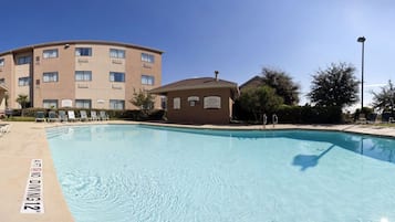 A swimming pool with lounge chairs, a small building, and a multi-story building in the background.
