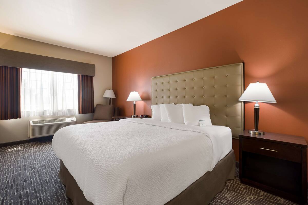 A hotel room with a large bed, two bedside tables with lamps, a window with curtains, and an air conditioning unit.
