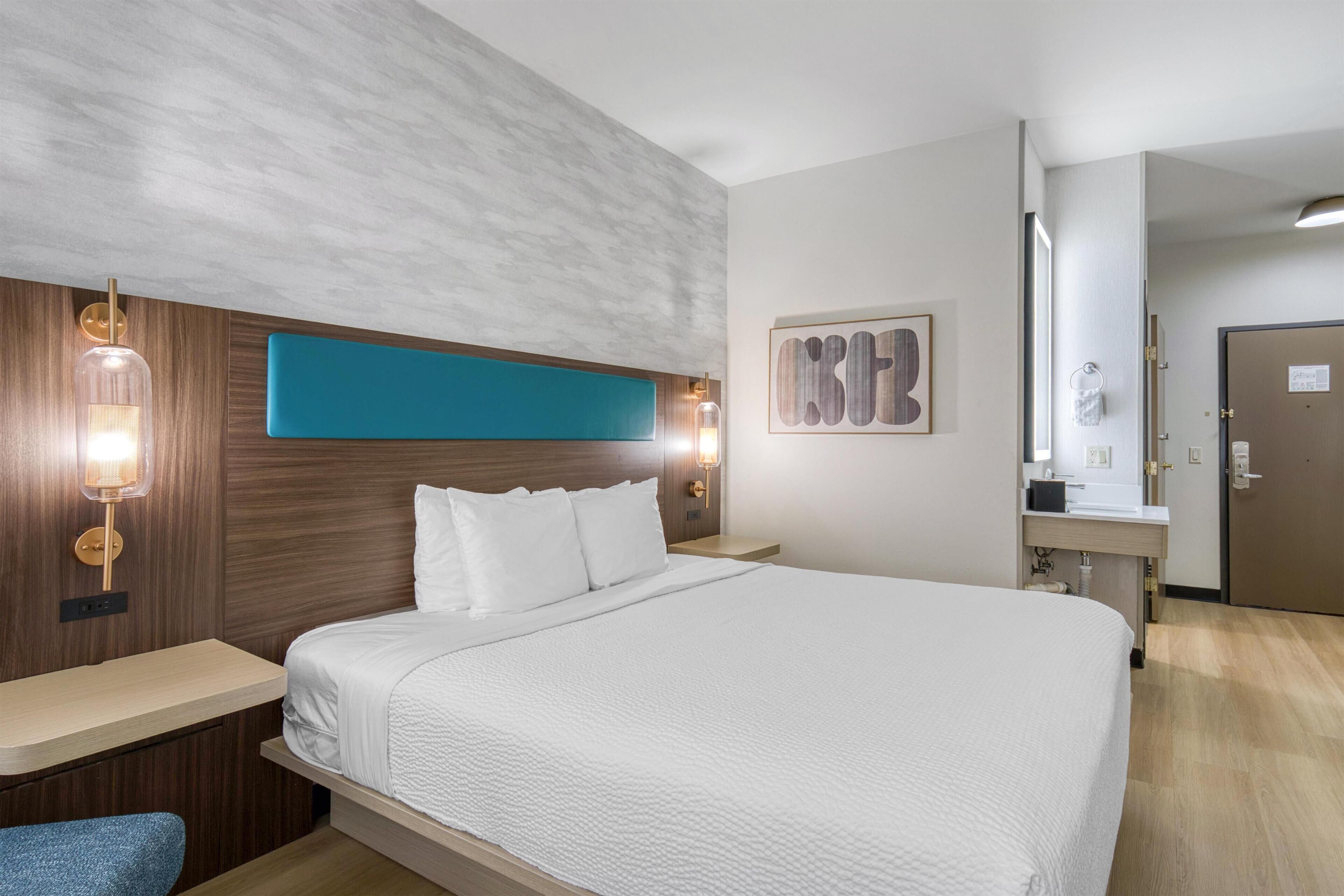 Ein Hotelzimmer mit einem großen Bett, einem Holzkopfstück, zwei Nachttischlampen, einem an der Wand hängenden Kunstwerk und einem durchgehenden Türspalt sichtbaren Badezimmer.