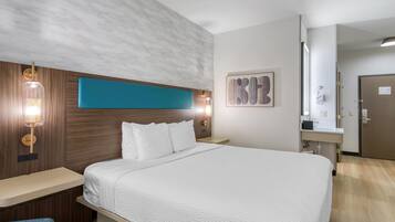 Ein Hotelzimmer mit einem großen Bett, einem Holzkopfstück, zwei Nachttischlampen, einem an der Wand hängenden Kunstwerk und einem durchgehenden Türspalt sichtbaren Badezimmer.