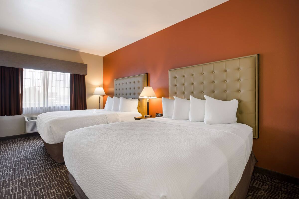 A hotel room with two beds, a large window with curtains, a wall heater, and a lamp on the nightstand.