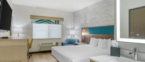 A hotel room with a bed, a desk, a chair, a TV, a window with blinds, a wall-mounted lamp, and a bathroom with a sink and mirror.