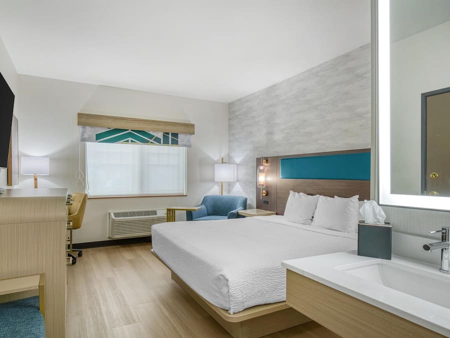 A hotel room with a bed, a desk, a chair, a TV, a window with blinds, a wall-mounted lamp, and a bathroom with a sink and mirror.