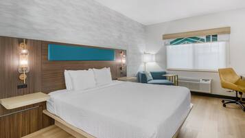 Una habitación de hotel con una cama grande, mesitas de noche, un escritorio, una silla, una ventana con persianas y una lámpara de pared.