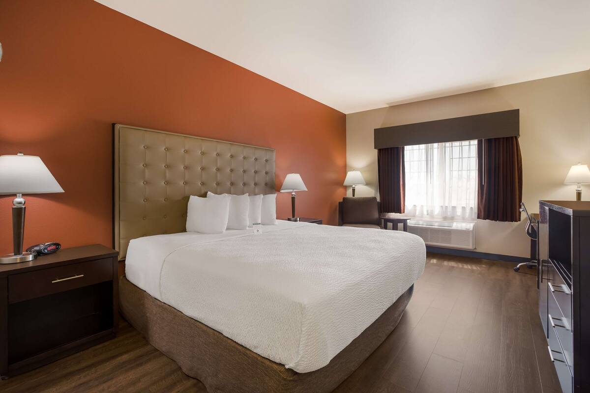 A hotel room with a large bed, two bedside tables with lamps, a desk with a chair, and a large window with curtains.