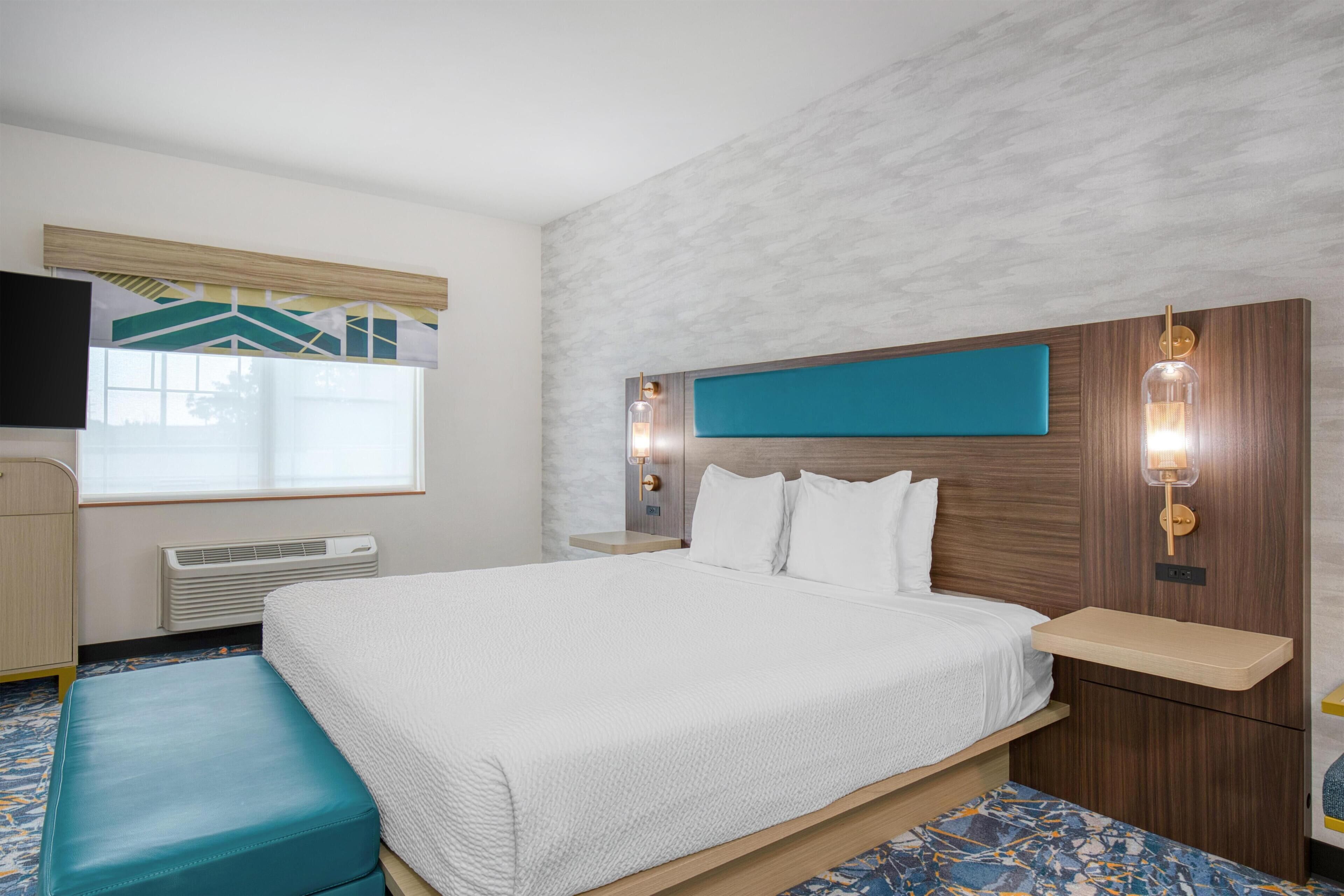 A hotel room with a bed, a bench, a TV, a window, and a wall-mounted light fixture.