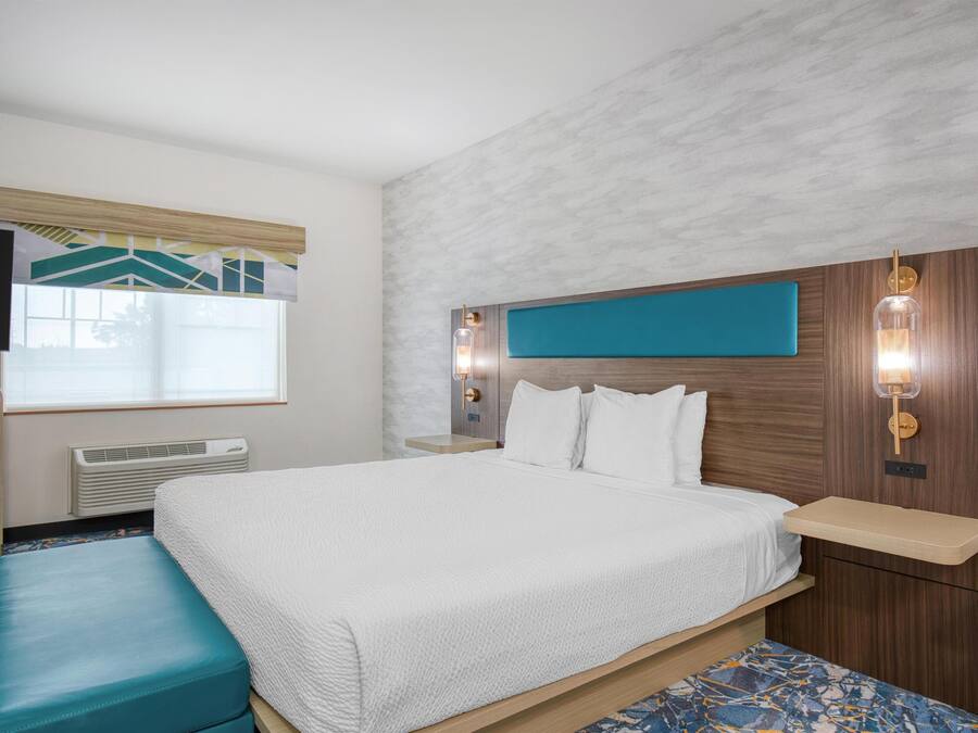 Habitación de hotel con cama, un banco, televisor, ventana y lámpara fijada a la pared.